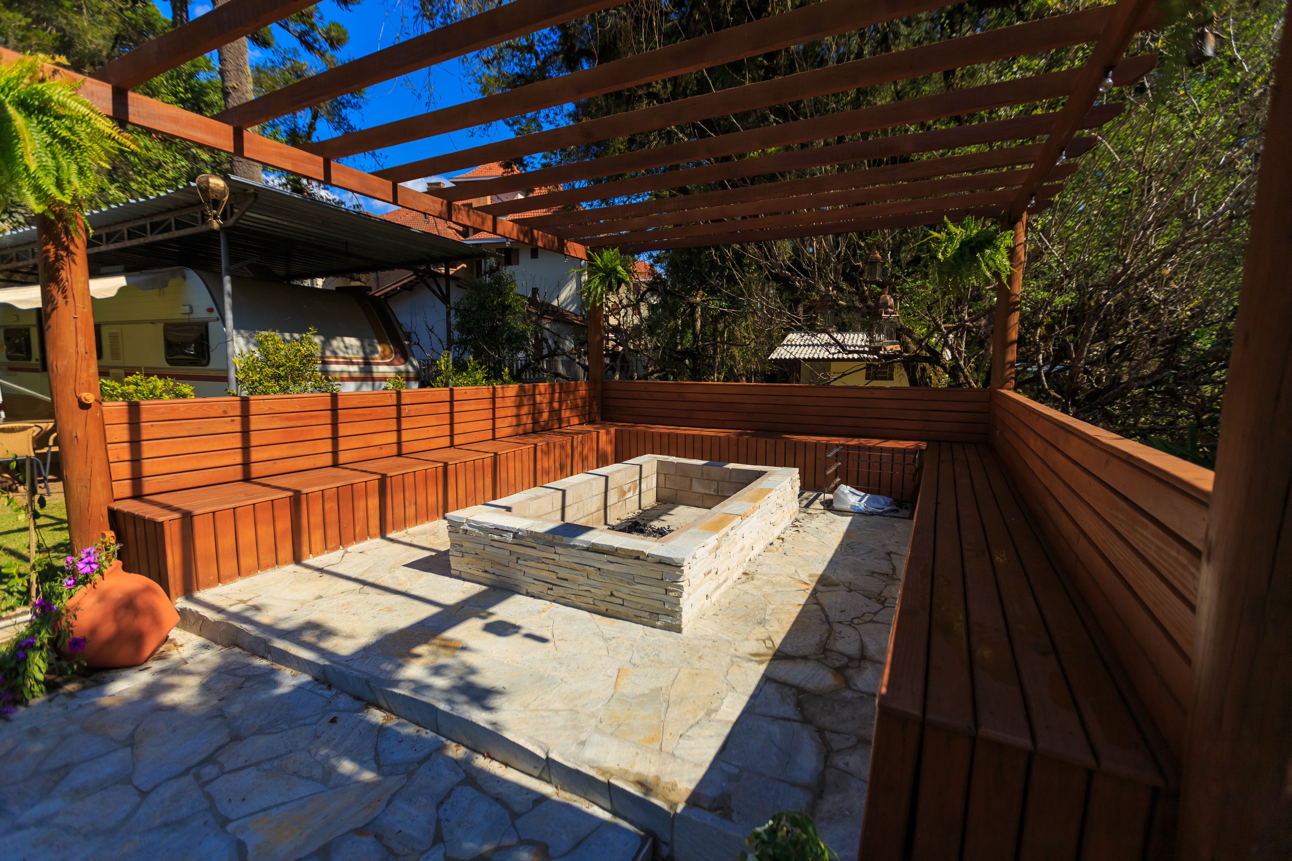 Stone fire pit and built-in wooden benches beneath a pergola create a cozy gathering space